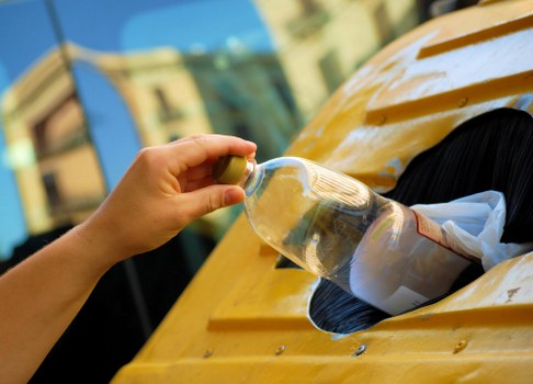 A close-up view of a person's hand holding a small yellow container and pouring water into a transparent plastic bottle, which is partially filled with liquid, on the yellow-painted surface of a waste collection vehicle or skip. The background shows an out-of-focus urban scene with buildings and clear daylight. This scene depicts waste management activity, possibly part of a rubbish removal or house clearance service in Garston, with attention to detail on the bottle and the vehicle's surface, reflecting the process of collecting or processing recyclable materials as part of the local rubbish removal services provided by House Clearance Garston.
