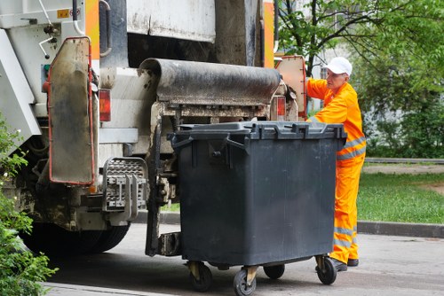 A waste collection operative wearing bright orange high-visibility workwear and a white safety helmet is positioned beside a large black wheeled rubbish bin on a suburban street during daytime. The bin has a textured plastic surface and is situated on the pavement, close to the side of a commercial waste vehicle with visible mechanical lifting components at the rear. The vehicle is beige with orange safety markings, and part of the lifting mechanism is engaged with the bin, suggesting an ongoing rubbish collection process. In the background, there are green trees and a grassy verge, indicating a residential or mixed urban area. The environment appears clean and orderly, with natural daylight illuminating the scene, emphasizing the textures of the bin's plastic surface and the reflective elements on the operative's safety gear. This scene reflects professional rubbish collection services, such as those offered by House Clearance Garston, serving local communities and supporting waste removal needs in the Garston and surrounding areas.