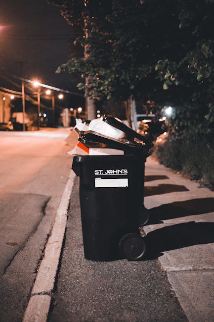 A black, cylindrical public rubbish bin situated on a cobblestone street curb, with various kinds of litter accumulated around its base including glass bottles, plastic bottles, crumpled paper, and fo